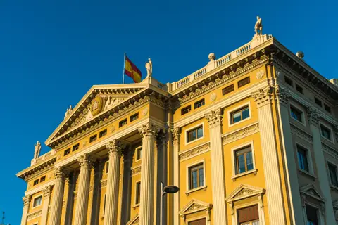 Façade néoclassique d'un bâtiment officiel surmonté du drapeau espagnol, sous un ciel bleu