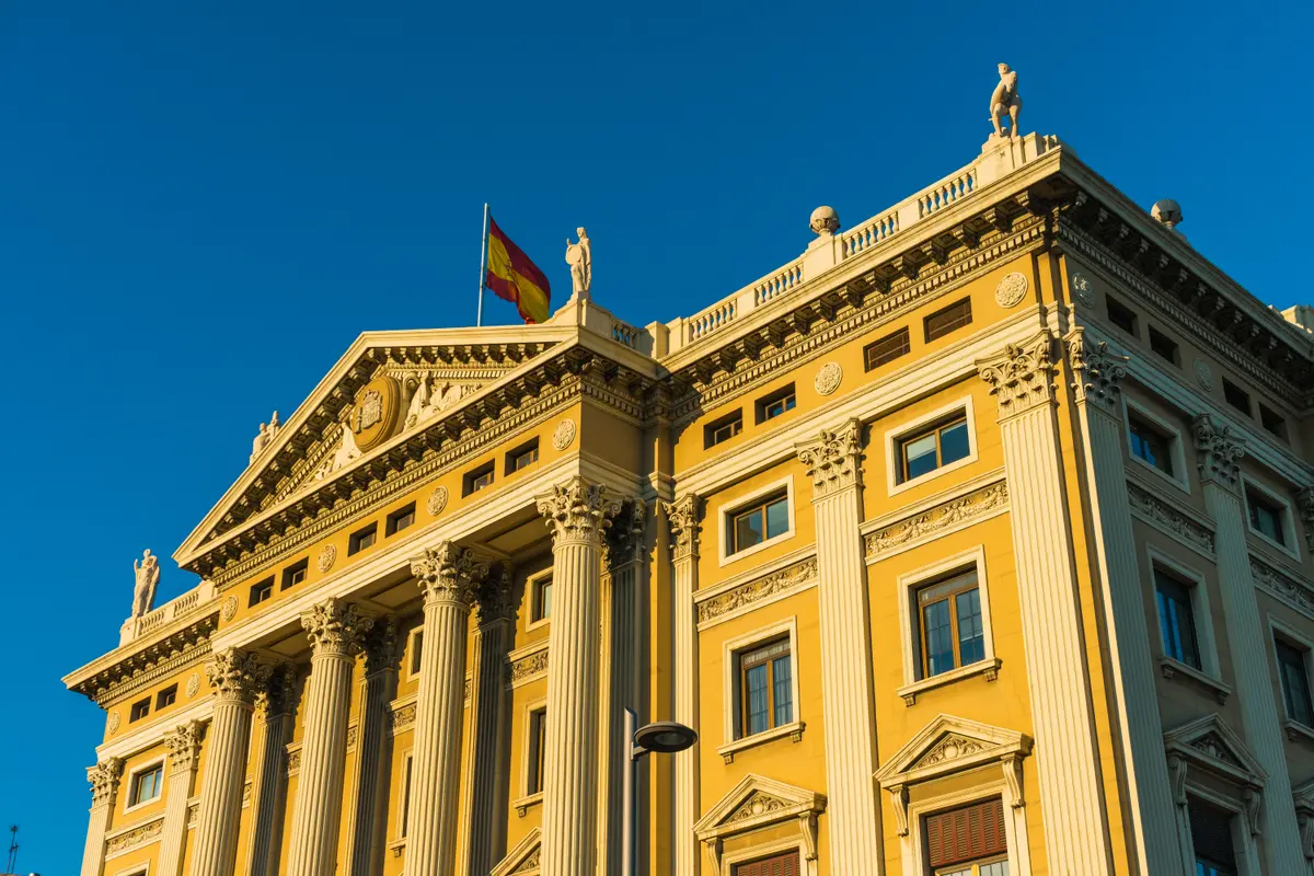 Façade néoclassique d'un bâtiment officiel surmonté du drapeau espagnol, sous un ciel bleu