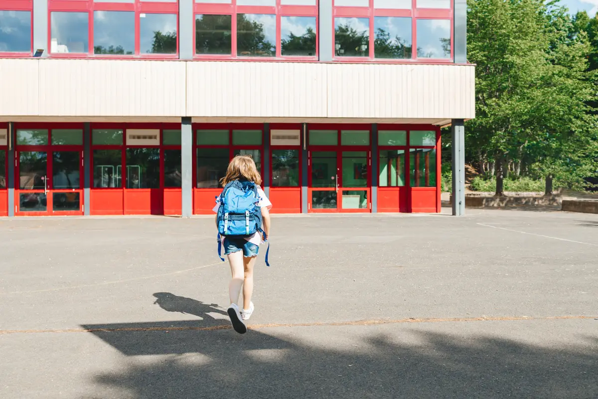 Fillette avec un sac à dos bleu courant dans la cour vers les portes rouges de son école