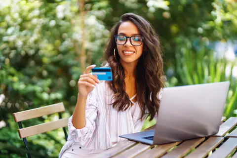 Femme tenant une carte bancaire devant son ordinateur portable, assise à une table de jardin